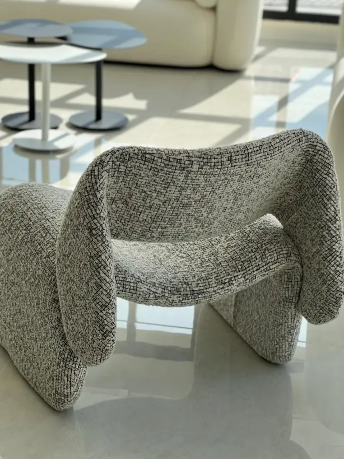 Contemporary sculptural chair with textured gray upholstery, set in a modern showroom with round nesting tables in the background.